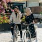 A man and woman in stylish attire biking together under sunny skies with floral surroundings.